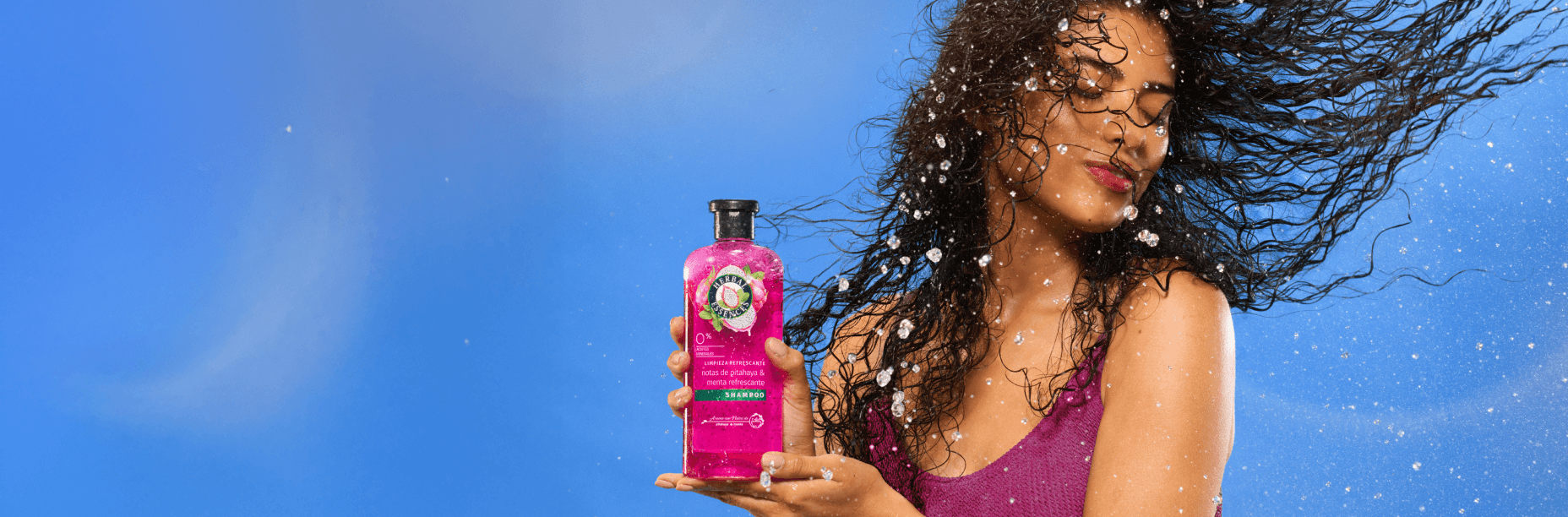 Mujer con cabello rizado y suelto sosteniendo productos Herbal Essences Pitahaya y Menta con texto Vibras Frescura