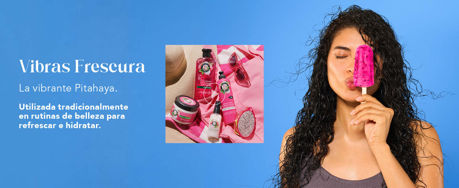 Vibras Frescura, la vibrante Pitahaya — colección Herbal Essences Pitahaya & Menta sobre una toalla rosa con sombrero y lentes, y una mujer de cabello rizado sosteniendo una paleta de pitahaya. Utilizada tradicionalmente en rutinas de belleza para refrescar e hidratar.