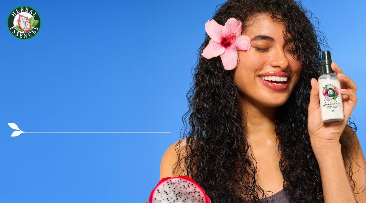 Mujer sonriendo con flor en el cabello sosteniendo Perfume Capilar Herbal Essences Pitahaya y Menta con texto Tamaño Ideal para acompañarte en tu bolso separa rocía sacude y disfruta